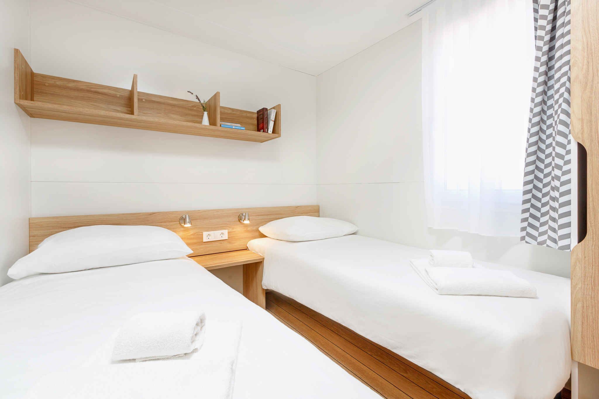 Une chambre d’hôtel avec deux lits, des têtes de lit en bois, une table de chevet avec une lampe, une étagère au-dessus des lits et une fenêtre avec un rideau.