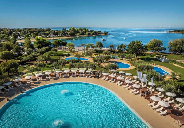 A resort with a large swimming pool surrounded by sun loungers and umbrellas, a marina with boats, and lush greenery.