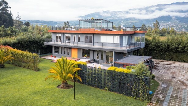 Une maison moderne à deux étages, dotée d’un balcon en verre, entourée d’une végétation luxuriante et offrant une vue sur un paysage montagneux.