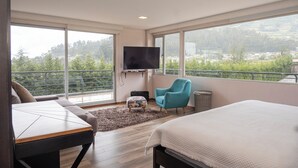 Une chambre moderne avec un grand lit, un canapé, un téléviseur fixé au mur et un balcon donnant sur un paysage verdoyant.