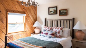 A cozy bedroom with a wooden ceiling, a bed with floral pillows, two bedside lamps, and a window with a view.