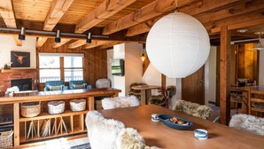 A rustic interior with a wooden dining table, fur-covered chairs, a hanging white paper lantern, and a brick fireplace.