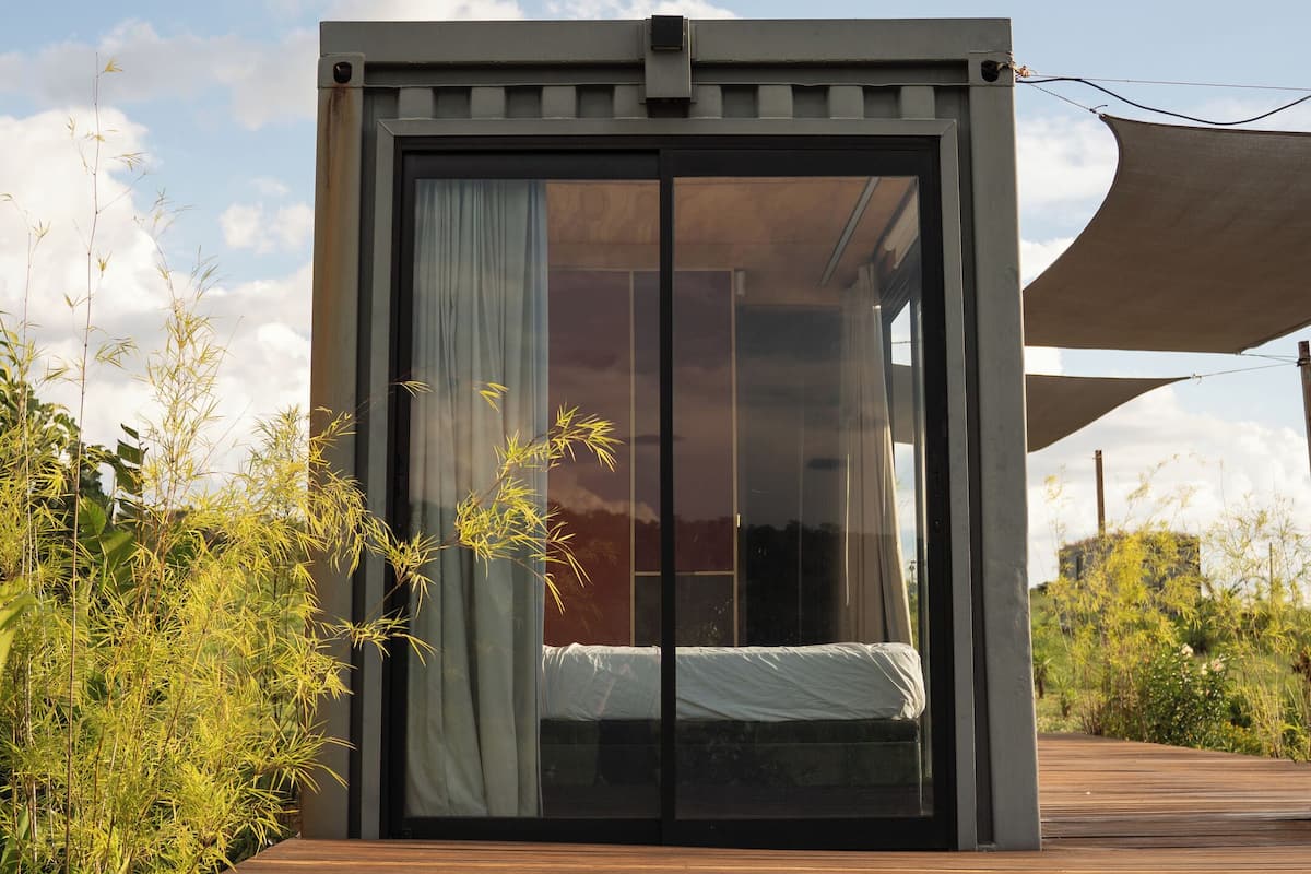 A modern, minimalist cabin with a large sliding glass door, a wooden deck, and lush greenery surrounding it.