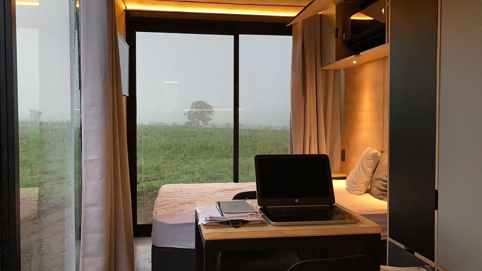 Un dormitorio con una cama, un escritorio con una computadora portátil y vistas a un campo verde a través de un ventanal grande.