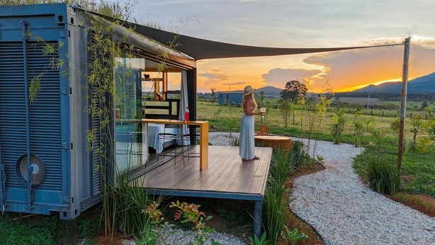 Een moderne hut met een terras, een eethoek en uitzicht op de ondergaande zon boven een bergachtig landschap.