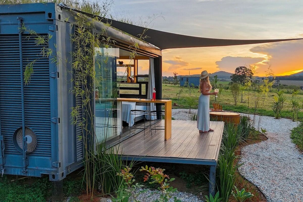 A modern cabin with a deck, a dining area, and a view of the sunset over a mountainous landscape.