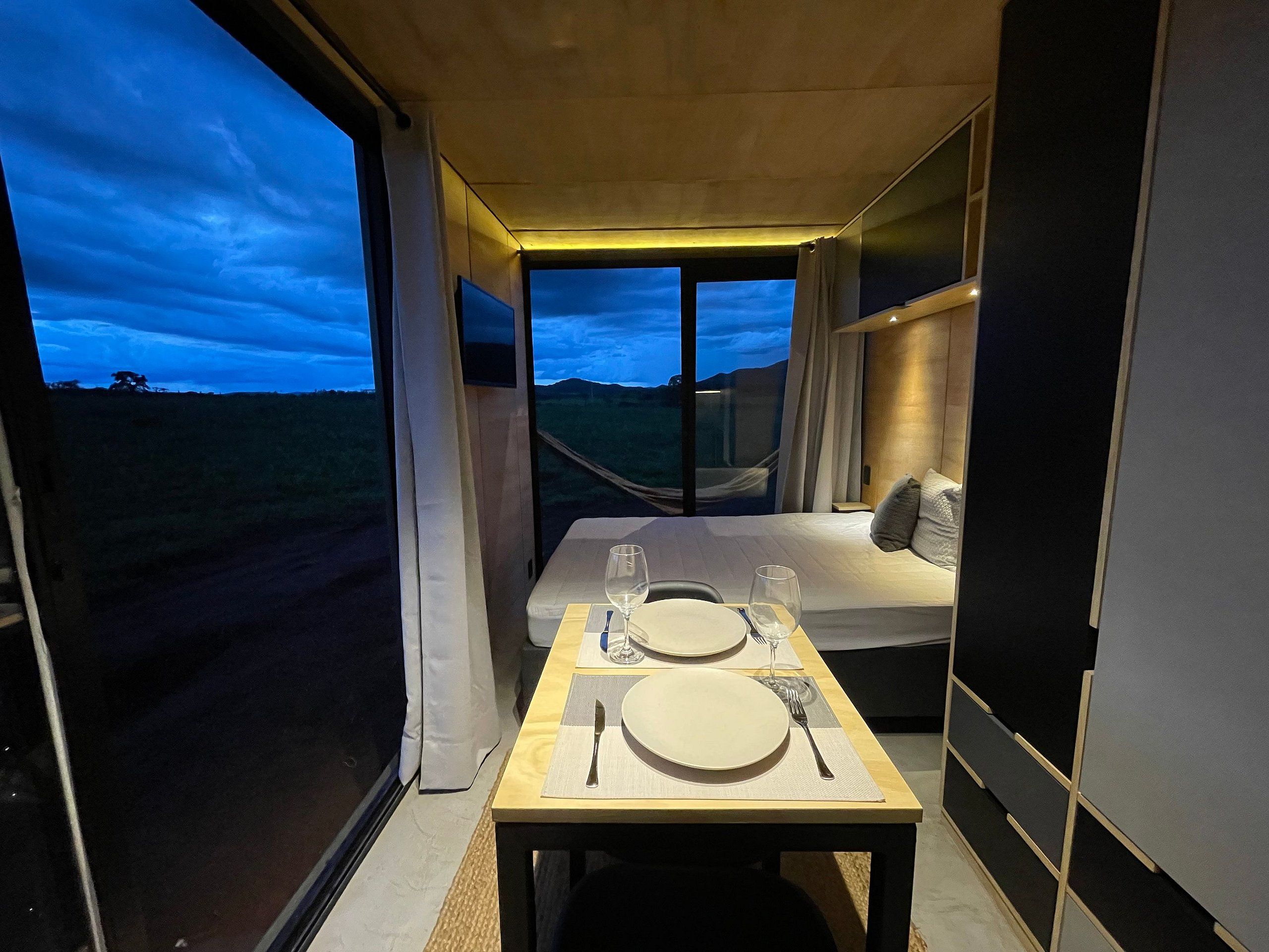 Quarto compacto com cama, mesa posta para dois e uma grande janela com vista para o exterior.