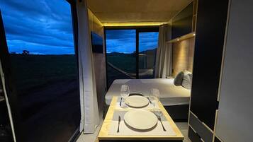 Quarto compacto com cama, mesa posta para dois e uma grande janela com vista para o exterior.