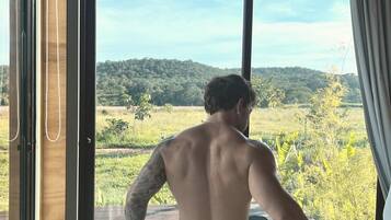 Uma pessoa sentada na cama, com vista para uma paisagem verde pela janela.