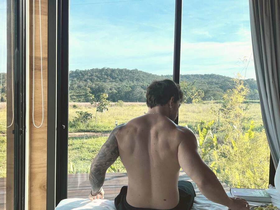A person sitting on a bed with a view of a green landscape through a window.