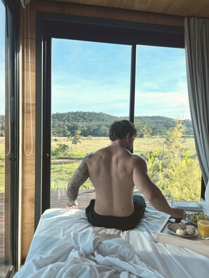 A person sitting on a bed with a view of a green landscape through a window. - Moriá Village (Pirenópolis)