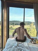 Una persona sentada en una cama con vista a un paisaje verde a través de una ventana.