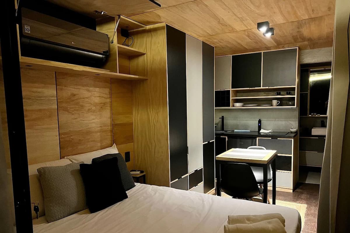 A compact hotel room with a bed, bedside table, and a kitchenette area.