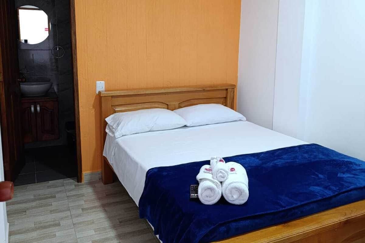 A hotel room with a wooden bed, white bedding, a blue blanket, and two rolled towels on the bed.