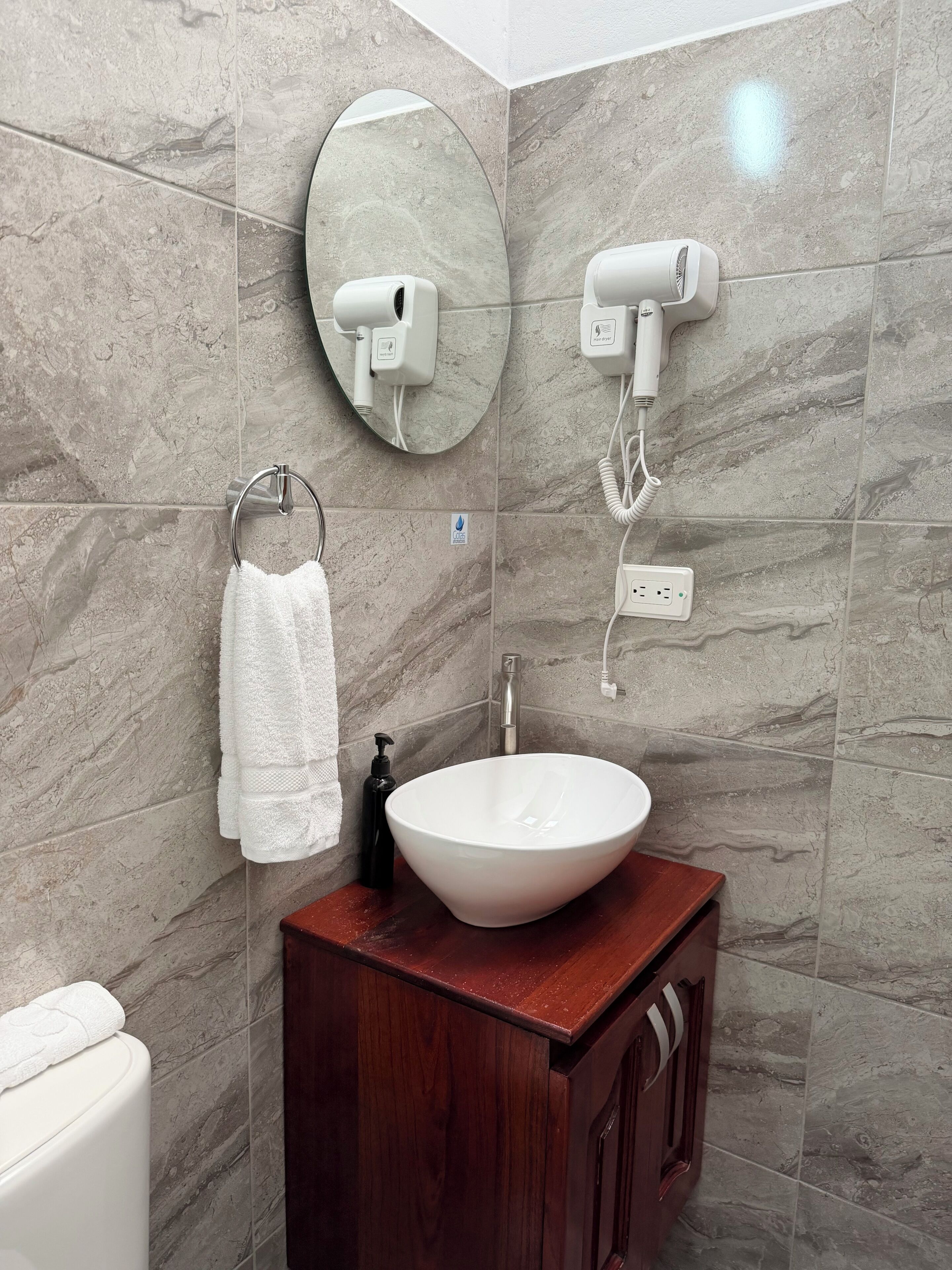 A bathroom with a marble wall, a round mirror, a white sink, a towel rack, and a hairdryer.