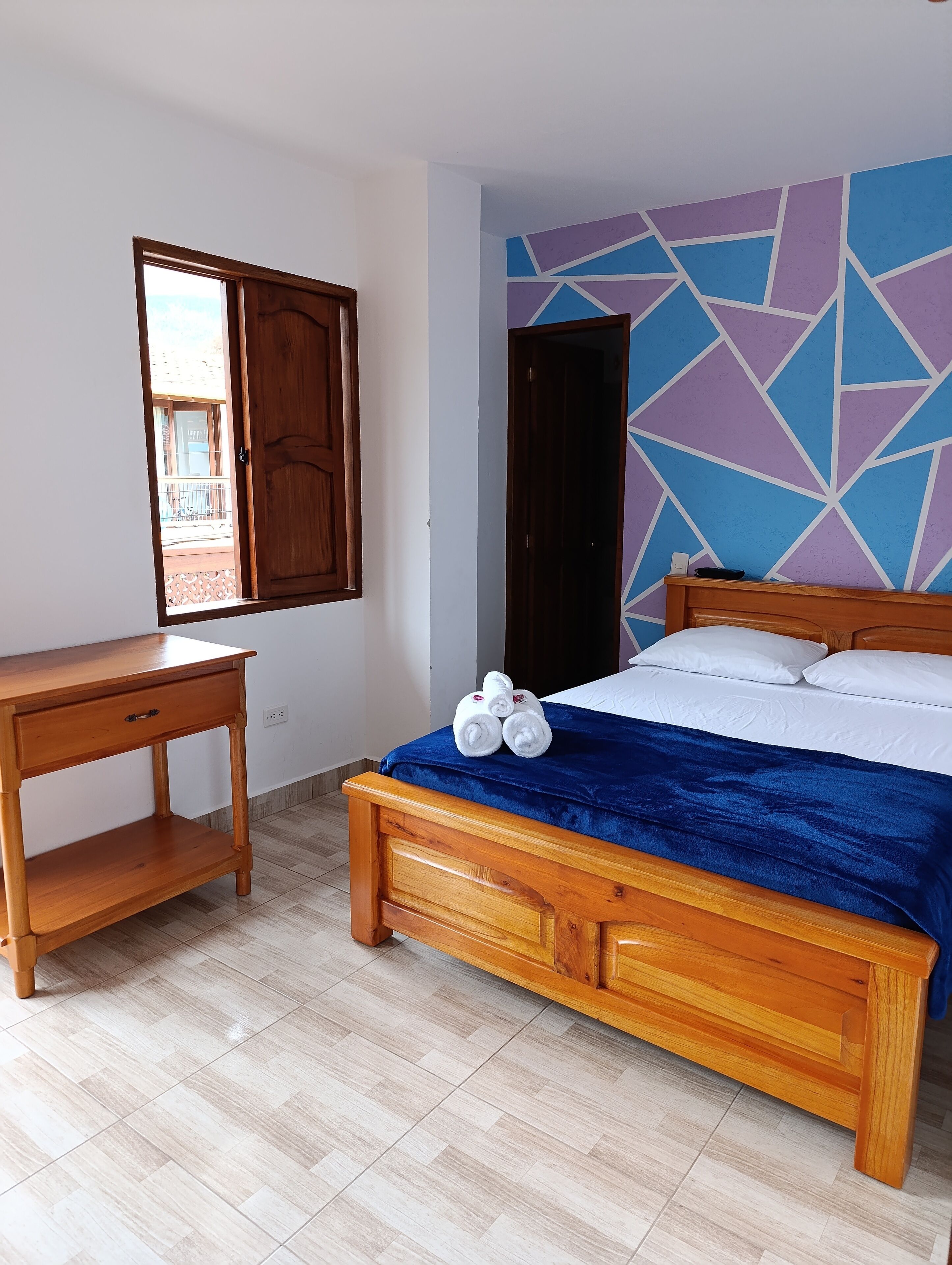 A bedroom with a wooden bed, a nightstand, a window with a view, and a geometric-patterned wall.