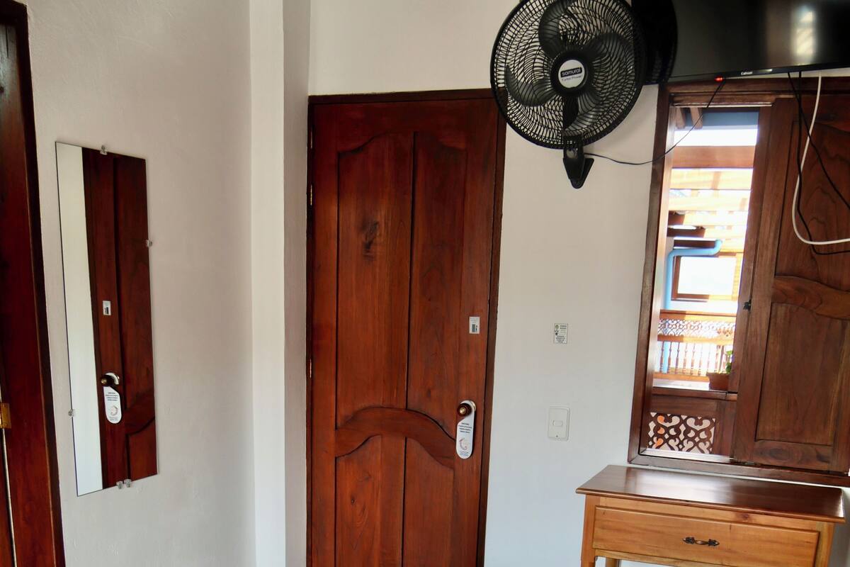 A hotel room with a bed, a wooden door, a small wooden table, and a fan.