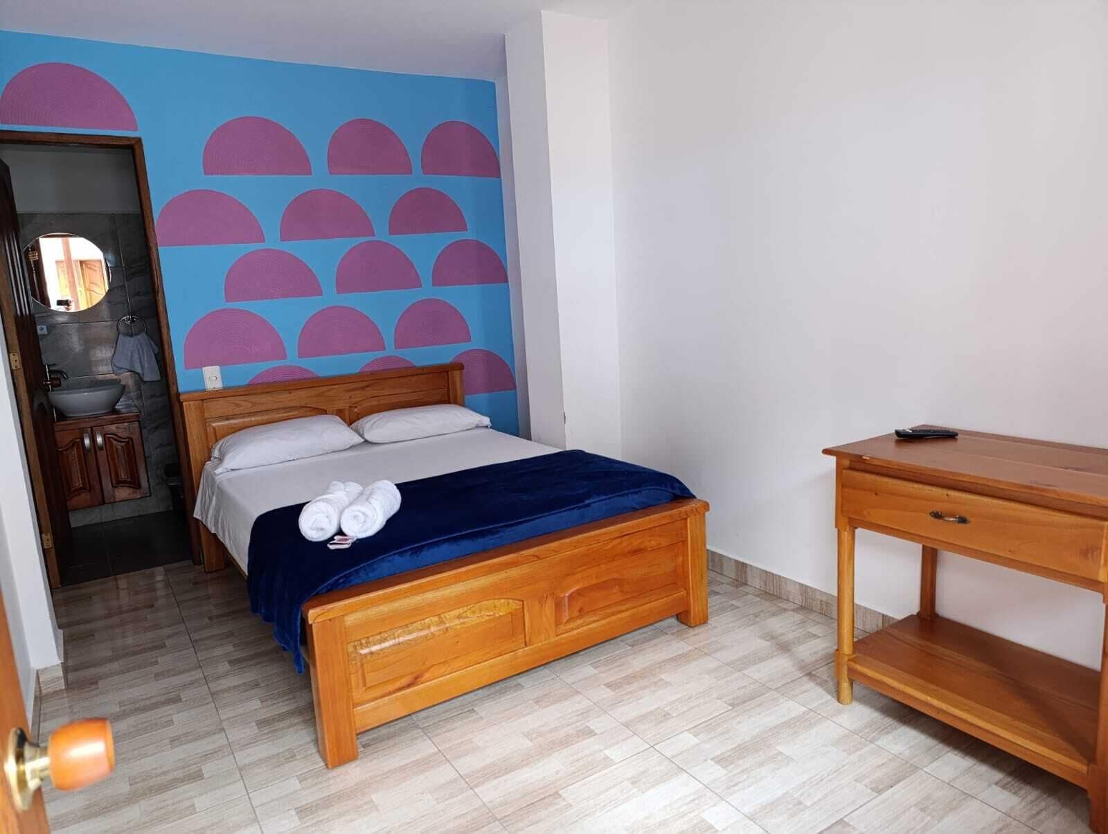 Ein Schlafzimmer mit einem hölzernen Bett, einer blauen Wand mit rosa Halbkreisen, einem Holztisch und einem Badezimmer mit Waschbecken und Spiegel.