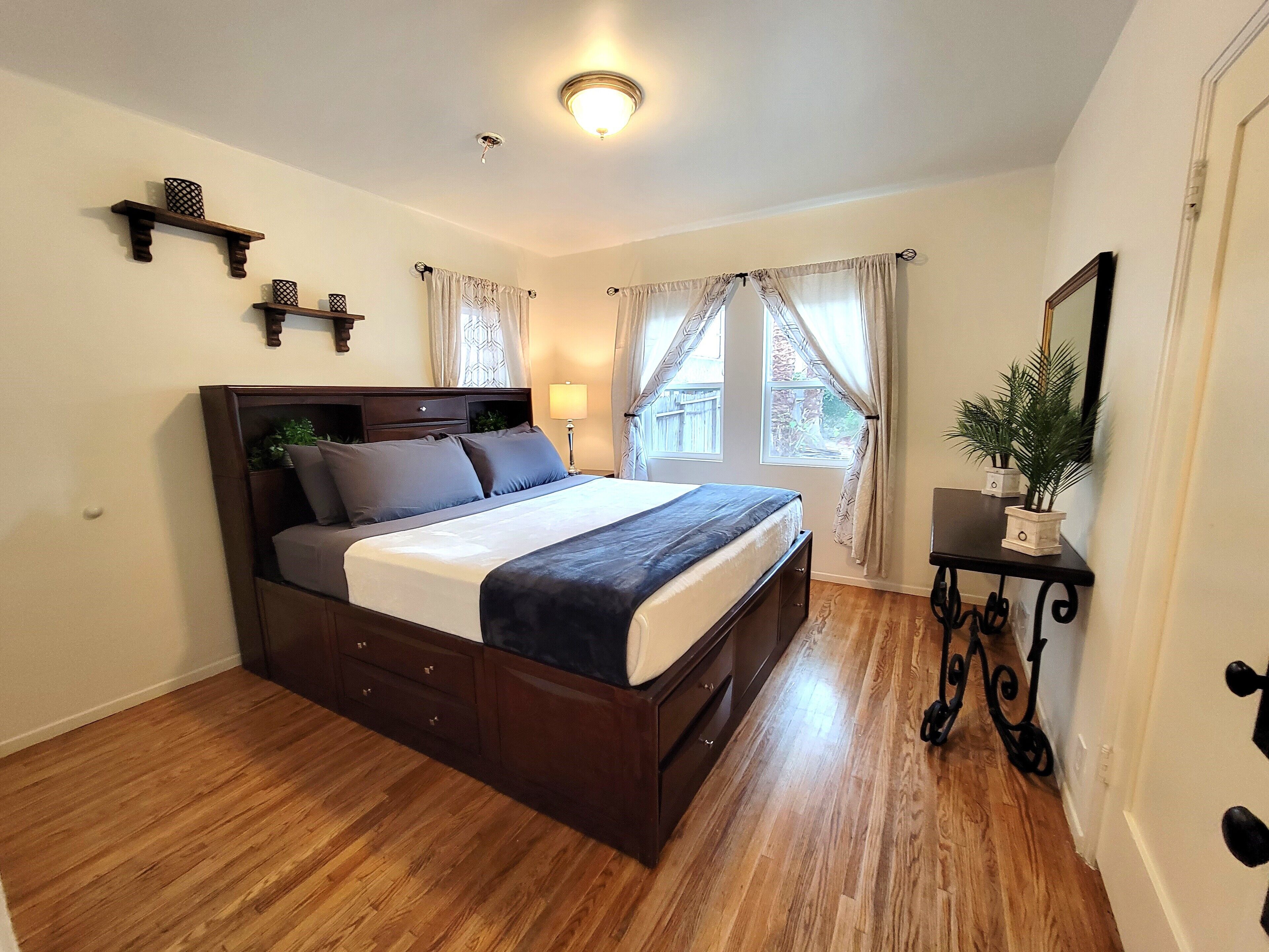A bedroom with a wooden bed, a nightstand with a lamp, a small table with a plant, and a window with curtains.