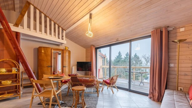 A spacious living area with a high ceiling, wooden flooring, a dining set, wicker furniture, and a large window offering a view of the outdoors.