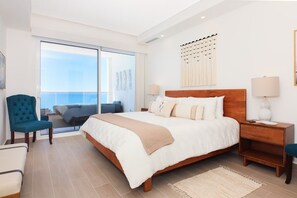 Quarto de hotel moderno com uma cama grande, mesa de cabeceira, cadeira e vista para a varanda.