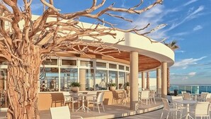 A seaside restaurant with outdoor seating, a wooden tree sculpture, and a view of the ocean.