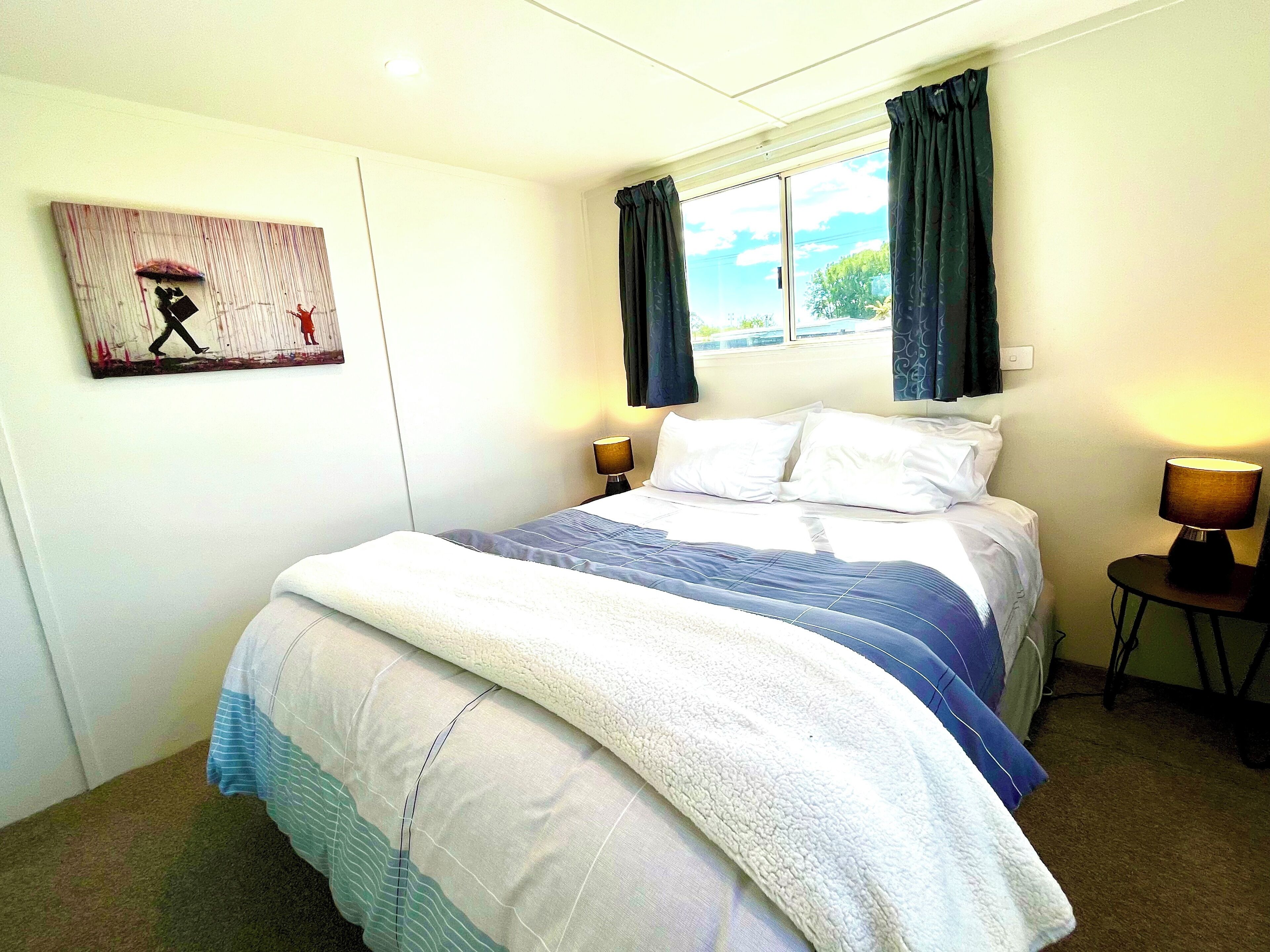 A neatly made bed with a blue and white comforter, two bedside lamps, a window with curtains, and a wall-mounted artwork.