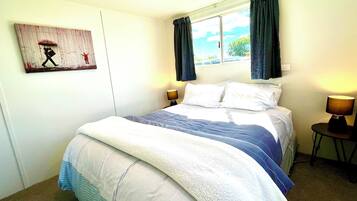 A neatly made bed with a blue and white comforter, two bedside lamps, a window with curtains, and a wall-mounted artwork.
