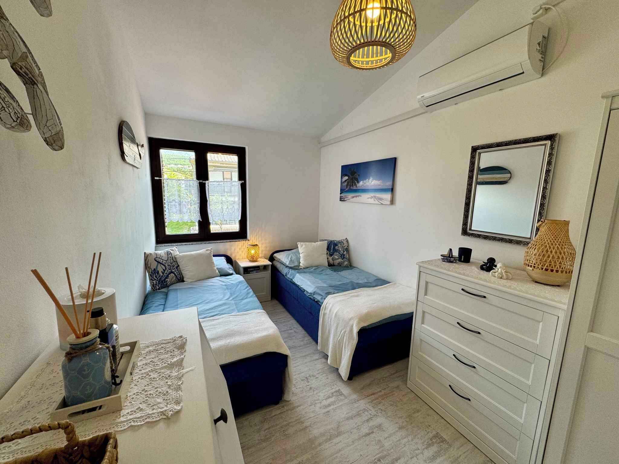 Un dormitorio con dos camas, un aparador, una ventana con cortinas de encaje, un cuadro y una lámpara colgante.