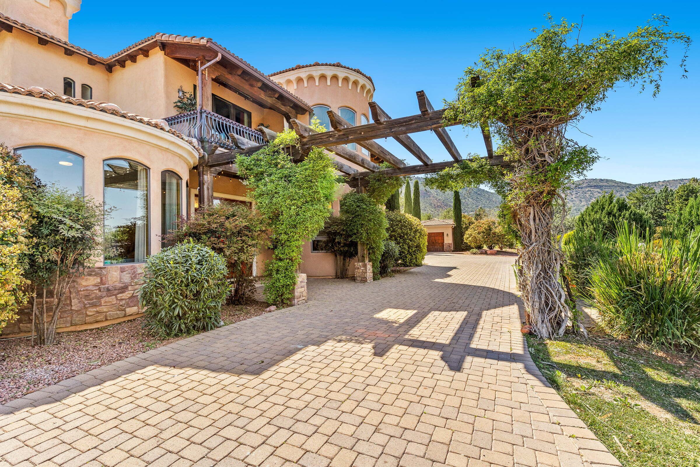 A house with a paved driveway, a wooden pergola, and a variety of greenery.
