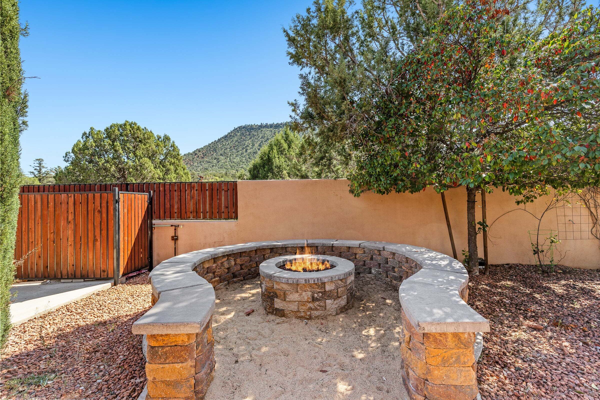 A circular stone fire pit surrounded by a low stone wall, with a wooden fence and a tree in the background.