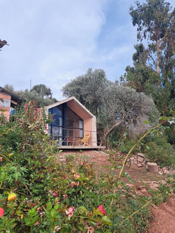 A small, single-story house with a sloped roof, a balcony with a chair, and a garden area with various plants and flowers. - Casa de Primo (Capachica)
