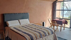 Un dormitorio con una cama grande, una mesita de noche de madera, una mesa con sillas y vistas amplias a través de ventanas grandes.