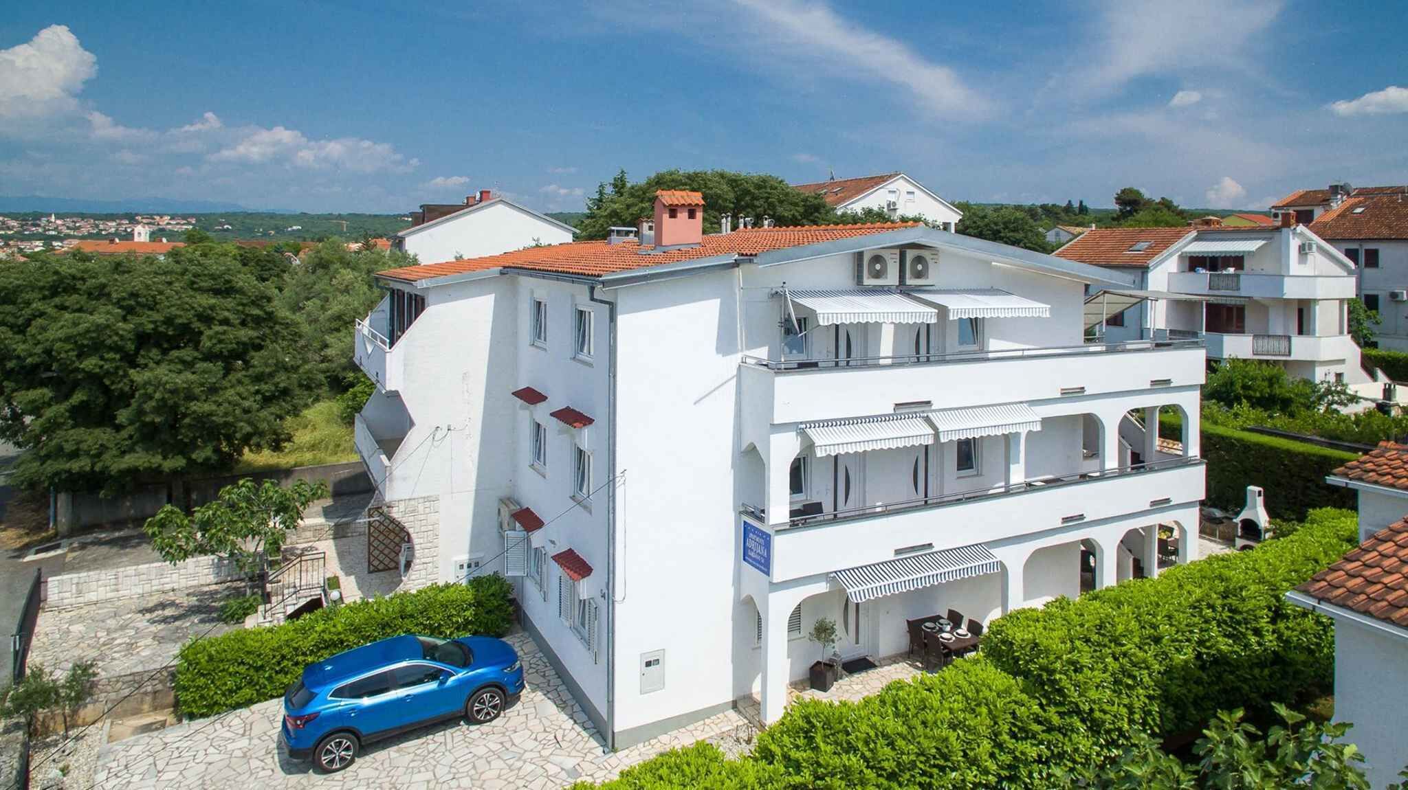 A multi-story white building with a red-tiled roof, a blue car parked in front, and a view of a town in the background.