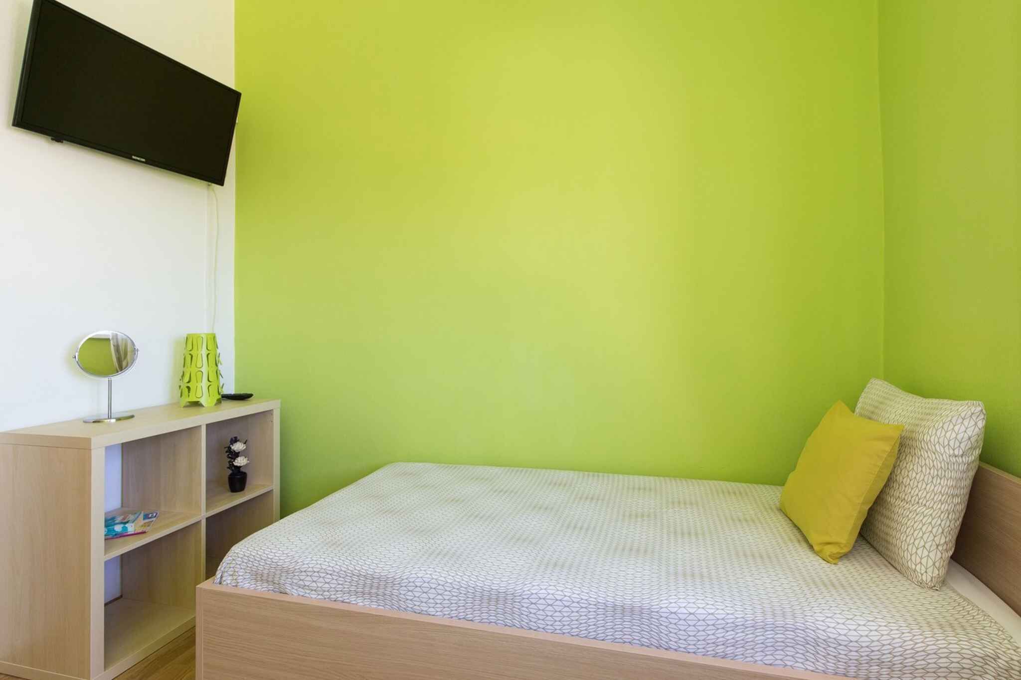 Una habitación con una cama, un televisor montado en la pared, una mesita de noche de madera y una pared verde.
