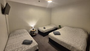 Una habitación pequeña y bien iluminada con tres camas, cada una con una colcha estampada y una toalla enrollada. A la derecha hay una pequeña mesa con una lámpara y una planta.