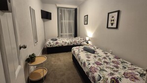 Un dormitorio pequeño y luminoso con dos camas individuales, una mesita de noche con lámpara, una pequeña mesa redonda con una planta y una ventana con cortinas de encaje.