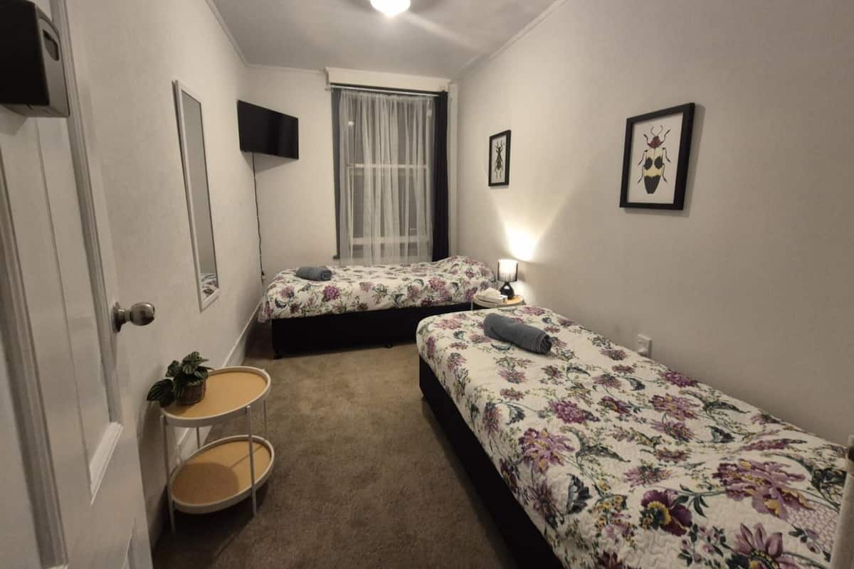 Ein kleines, gut beleuchtetes Schlafzimmer mit zwei Einzelbetten, einem Nachttisch mit Lampe, einem kleinen runden Tisch mit Pflanze und einem Fenster mit durchsichtigen Vorhängen.