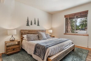 A bedroom with a wooden headboard, a bed with a grey quilt, two bedside tables with lamps, a small wooden nightstand, and a window with a view of trees and a car.