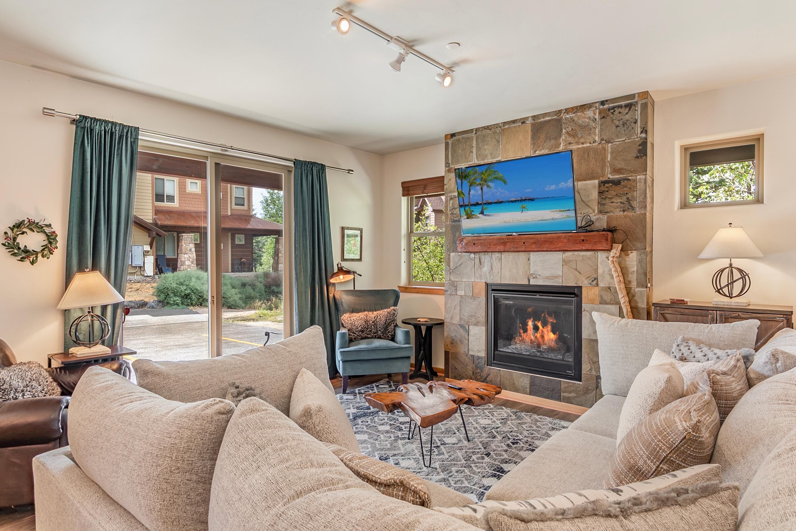 A living room with a fireplace, a television mounted on a stone wall, and a view of the outdoors through a sliding glass door.