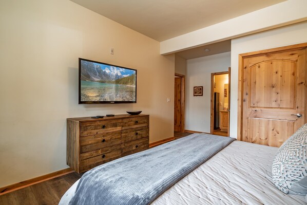 A bedroom with a bed, a wooden dresser, a television mounted on the wall, and a door leading to another room.