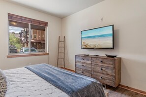 A bedroom with a bed, a dresser, a television, and a window with a view of buildings and parked cars.