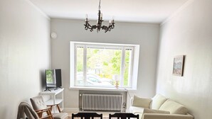 Un coin repas avec une table et des chaises, un radiateur, un canapé, un téléviseur et une fenêtre donnant sur des arbres et des voitures garées.