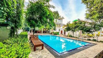A rectangular swimming pool with a tiled deck, surrounded by lush greenery and wooden lounge chairs.