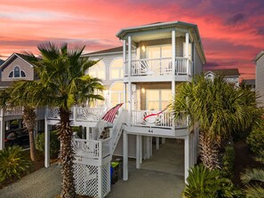 A two-story house with a balcony, palm trees, and a sunset sky. - 5BR/5BA Coastal Family Home: Large Deck, Community Pool, 5-Min Walk to Beach (Ocean Isle Beach)