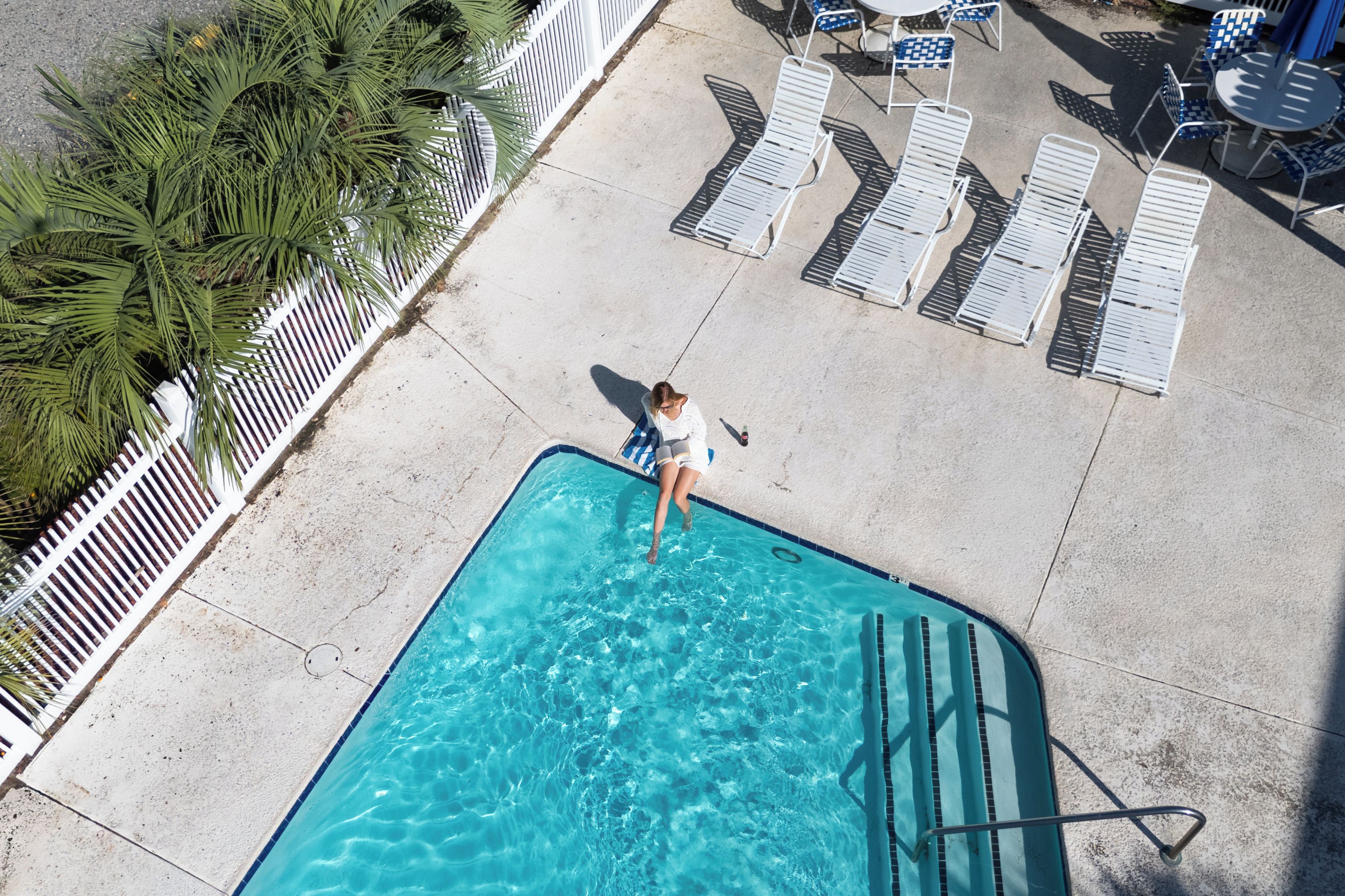 A swimming pool with a person standing in the water, surrounded by lounge chairs and a palm tree.