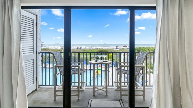 A balcony with a view of the beach, a pool, and two chairs.