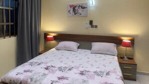 Un dormitorio con cama, ventilador de techo, equipo de aire acondicionado, mesita de noche con lámpara y una obra de arte en la pared.