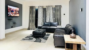 A modern living room with a flat-screen TV, a black leather sofa, a wooden coffee table, and a ceiling fan. - T-Roys Apartments (Accra)
