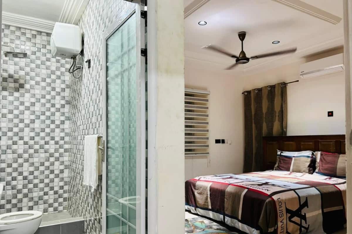 La salle de bain est dotée d’un mur recouvert de carreaux à motifs, d’une cabine de douche en verre et de toilettes. La chambre comprend un lit avec une courtepointe, un ventilateur de plafond et une fenêtre avec des rideaux.